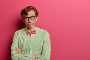 Bored adult man geek sighs and looks thoughfully aside, keeps arms folded, wears spectacles and...