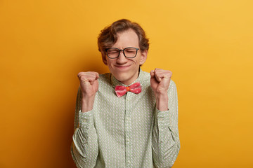 Ecstatic male nerd raises clenched fists, believes in success, closes eyes and awaits announcement of results, participates in contetst, hopes to win, expresses impatience, dressed formally. - obrazy, fototapety, plakaty