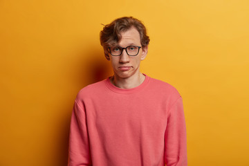 Bored unimpressed male student looks seriously at camera, has fatigue expression, wears optical glasses and pink jumper, sighs from tiredness, isolated on yellow background. Face expressions