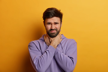 Displeased man suffocates because of painful strangle in throat, touches neck, looks dissatisfied at camera, has sore throat after catching cold, dressed casually, poses over yellow background