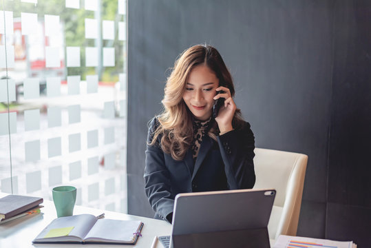 Happy Young Asian Woman Talking On The Mobile Phone And Smiling While Sitting At Her Working Place In Office.