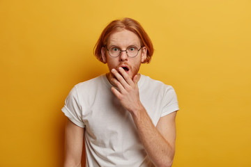 Stunned redhead man reacts on seasonal discounts, gazes with stupor at camera, covers mouth, wears spectacles and white t shirt, isolated on yellow background, forgot something. Omg expression