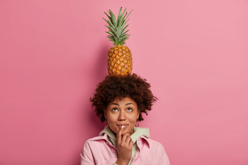 Photo of curious dark skinned African American woman carries pineapple on head, looks above, dressed in fashionable clothes, poses against pink background, has positive mood, plays with exotic food