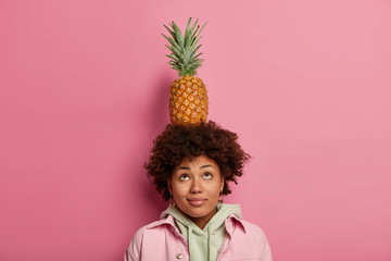 Pretty curly woman looks above, keeps balance with pineapple on head, concentrated above, has dark skin and curly hair, dressed casually, isolated on pink wall. Lovely female carries tropical fruit