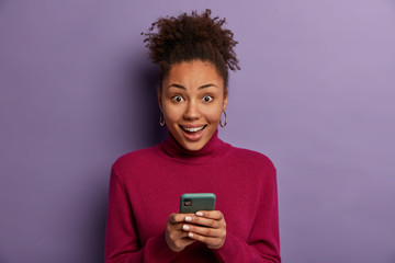 Happy surprised curly haired woman receives good news from friend, holds mobile phone, sends text...