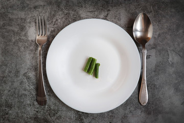 A round white plate with some food and a metal fork with a spoon on a gray cement background. The concept of anorexia, weight loss, starvation, lack of food. Improper nutrition. Diet.