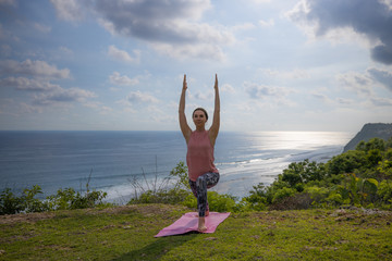 Young woman practicing yoga, standing in Virabhadrasana pose with raising arms. Outdoor yoga on the cliff. Warrior pose. Bali, Indonesia