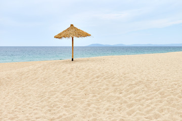 Single straw umbrella on golden sandy beach against blue sky and sea line; seascape for a creative story about the loneliness or sadness on the beach