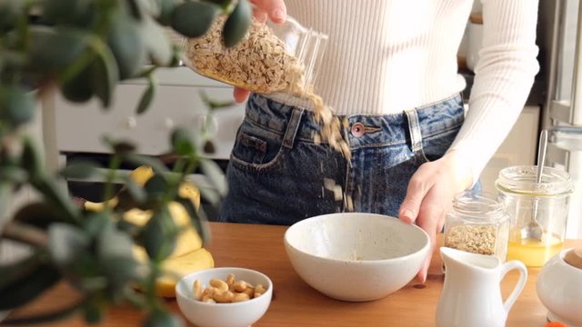 Woman preparing breakfast oatmeal porridge. Oat flakes falling in bowl. Concept of healthy eating, healthy lifestyle, dieting, vegan vegetarian lifestyle. Female prepares food at sunny kitchen