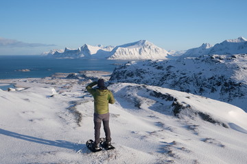 A man with a snoweshoes is looking from a viewpoint  to beautiful coast of  Lofoten islands.  Very popular touristic area for outdoor activities as hiking, skiing, surfing. 