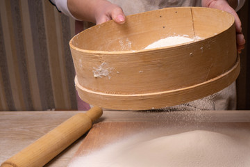 A woman sifts flour through a sieve. Plywood cutting board, wooden flour sieve and wooden rolling pin - tools for making dough.