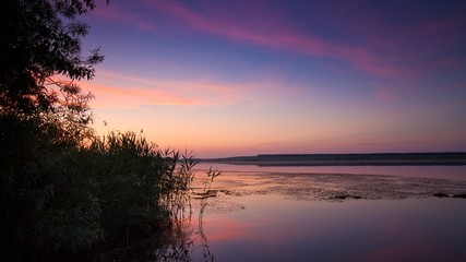 Fototapeta na wymiar romantic twilight with tender pink and purple clouds in deep blue sky, bulrush thickets and willow branches at still water surface of a countryside lake and reflections, peaceful summer sunset