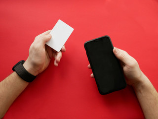 Hand with smart watch holding white card and smartphone on red background. Cashless payment, online shopping.