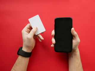 Hand with smart watch holding white card and smartphone on red background. Cashless payment, online shopping.