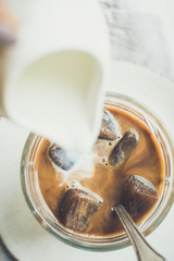 Pouring milk from a jug to a glass of espresso ice cubes