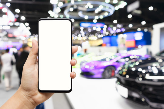 Mock up mobile phone. Hand holding smartphone with  Blurred image of public event exhibition hall showing cars and automobiles. buying new car concept. Mockup for your advertisement.