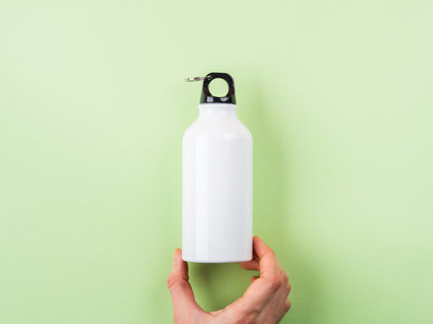 Zero waste concept - white metal reusable water bottle held by female hand. Flat lay on green pastel background