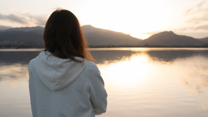 Pensive young woman with long hair seen from behind looks at the lake at sunset