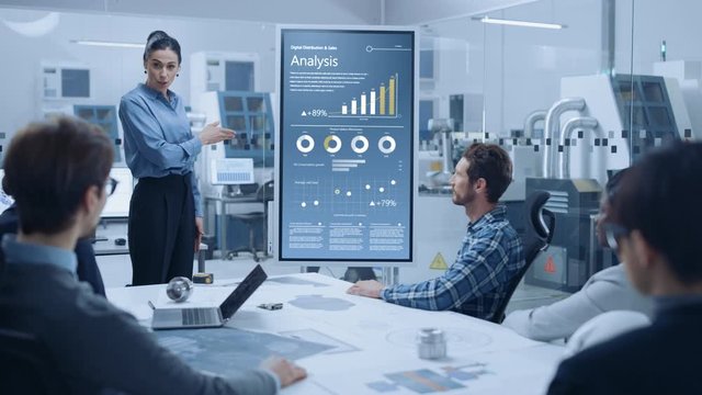 Modern Industrial Factory Meeting: Confident Female Engineer Uses Interactive Whiteboard, Makes Report to a Group of Engineers, Managers Talks, and Shows Statistics, Growth and Analysis Information