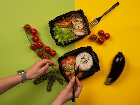 Woman Hands With A Fork Reaching For Food In Takeaway Box: Rice With Fish In Sauce, Chicken With Buckwheat, Vegetables And Greens. Two Meals With Cherry Tomatoes, Eggplant And Rosemary Flatlay.