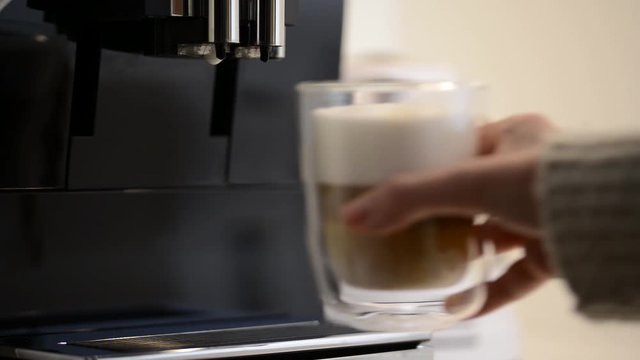 Young woman hand taking a clear glass cup of fresh latte macchiato coffee from modern coffee machine.