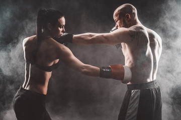Fototapeta na wymiar Shirtless Woman exercising with trainer at boxing and self defense lesson, studio, dark background. Female and male fight.