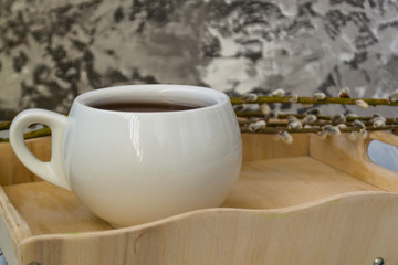 A willow twig with buds and a white cup of tea stands on a wooden tray. The concept of morning, breakfast