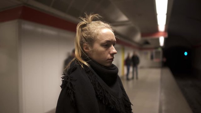 Handheld middle shot of blond woman in a black coat with a black scarf standing on metro platform in Budapest. Woman alone waiting for a train in a Budapest metro station