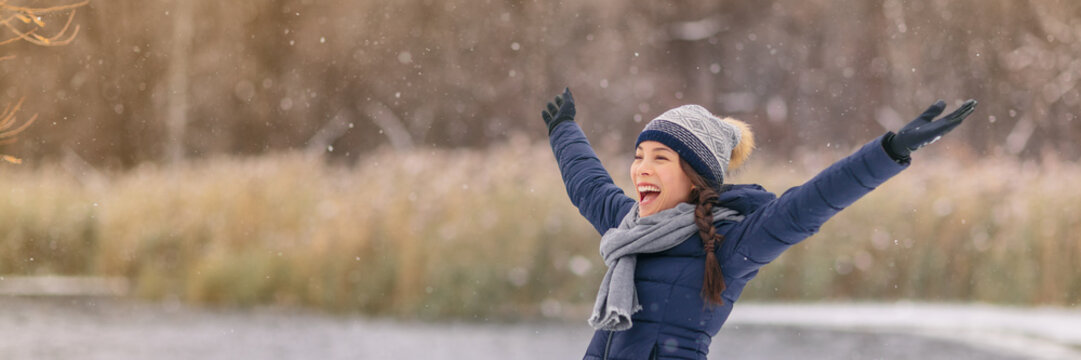 Winter Snow Happy Woman With Open Arms In Fun Enjoying Snow Fall Falling Snowflakes Wearing Cold Weather Scarf, Hat, Gloves Warm Jacket Banner Panorama. Girl Walking Outdoor Nature Woods Background.