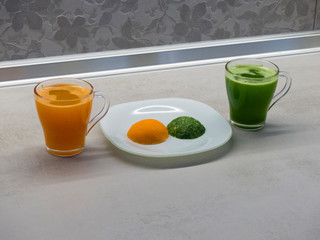 two cups, one with a carrot smoothie and the second with a cucumber smoothie next to the carrot and cucumber pulp on a white hemispherical plate