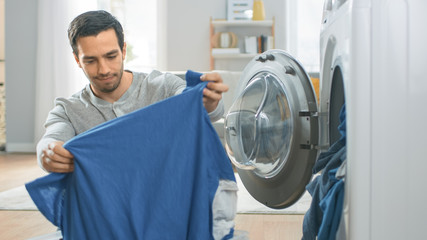 Handsome Smiling Young Man in Grey Jeans and Shirt Sits in Front of a Washing Machine at Home. He...