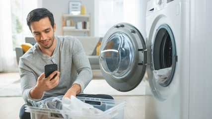 Handsome Smiling Young Man in Grey Jeans and Coat Sits in Front of a Washing Machine and Uses His...