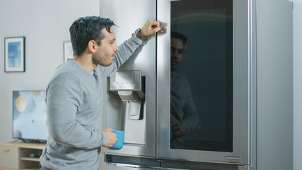 Handsome Young Man is Standing Next to a Refrigerator While Drinking His Morning Coffee. He is Checking a To Do List on a Smart Fridge at Home. Kitchen is Bright and Cozy.