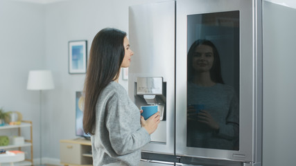 Beautiful Young Girl Stands Next to a Refrigerator While Drinking Her Morning Coffee. She is Checking the Weather Forecast and a To Do List on a Smart Fridge at Home. Kitchen is Bright and Cozy.