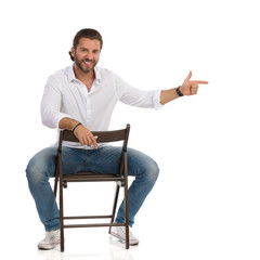 Handsome Smiling Man Is Sitting On A Chair, Looking At Camera And Directing.