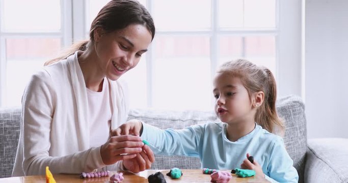 Female babysitter helping cute kid girl sculpting playdough together