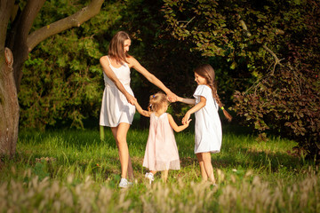 Naklejka na ściany i meble family mom and two daughters in light dresses hold hands and dance in a green park