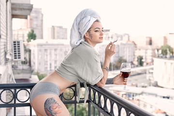 Home Leisure. Young woman in underwear and towel standing on balcony with glass of wine smoking...