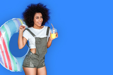 Swimming time. girl wearing denim overall standing isolated on blue with swim ring drinking smoothie joyful