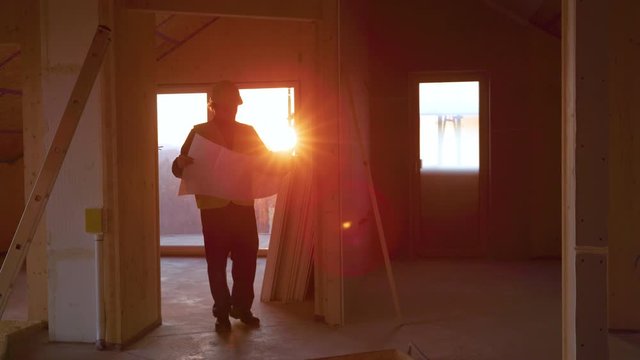 SLOW MOTION, LENS FLARE, DOF: Sunset illuminates the hallways as static engineer walks around the house with the floor plans in hand. Architect walks around the prefabricated house under construction