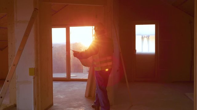 SLOW MOTION, LENS FLARE, DOF: Static engineer walks around a prefabricated house under construction with the floor plans in his hand. Project manager walks around a sunlit CLT house under construction