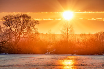 Orange winter sunrise over the frozen river and bare trees