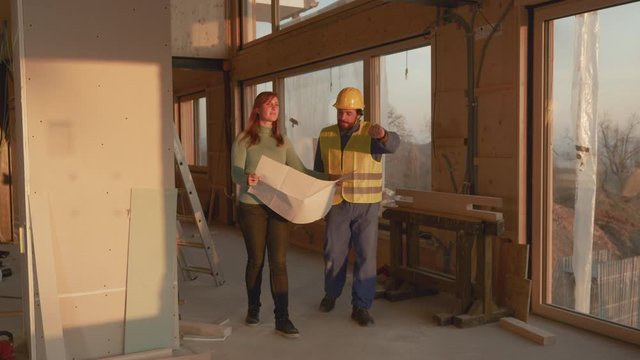 Architect holding his plans gives a young Caucasian woman a tour of her beautiful wooden house under construction. Future home owner and contract walk around a construction site and discuss the plans.