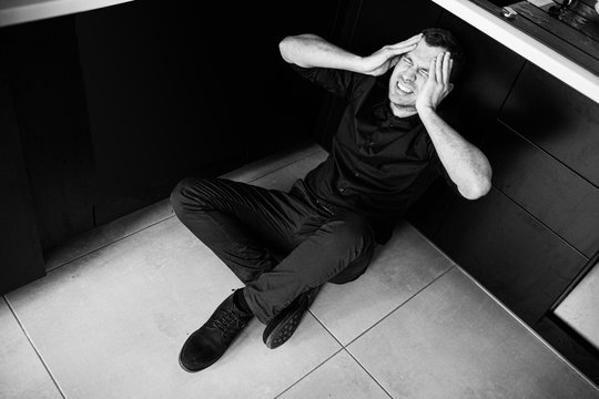 Black and white picture of young man in black clothes sitting separated alone in kitchen and scream. Hold hands on head and try to calm down. Disorder and pain. Desparation.