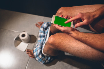 Man sit on pot in toilet or rest room. Guy hold phone with green screen and play with it. Point...