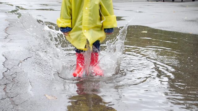 Slow motion video of little child in red rubber boots splashing water and jumping in big puddle after rain on street