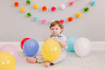Fototapeta na wymiar Funny Caucasian baby boy celebrating first birthday. Child kid toddler sitting on floor with colorful balloons. Celebration of event or party indoors at home. Happy birthday lifestyle concept.
