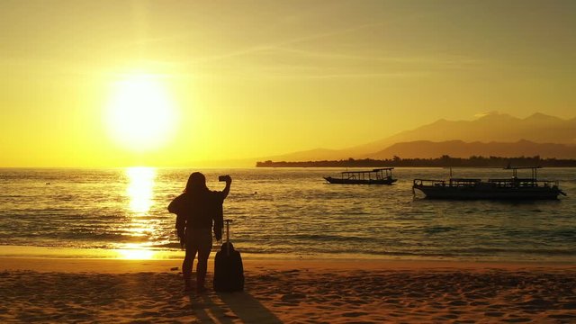 Tourist girl with travel bag taking pictures on shore of tropical island at sunset with yellow sun and sky reflecting on sea with anchored boats
