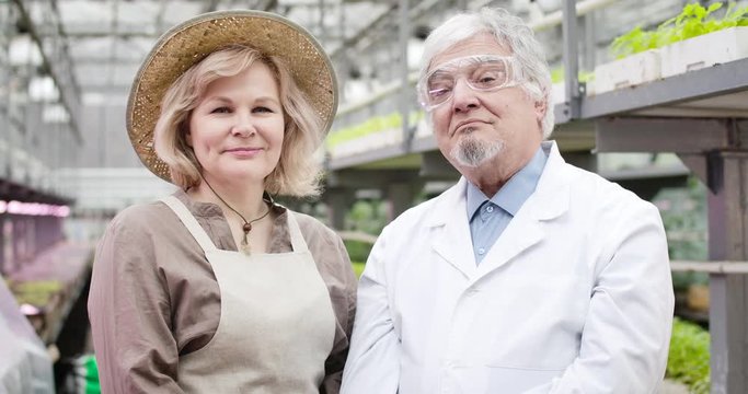 Portrait of senior Caucasian woman in straw hat and man in protective eyeglasses looking at camera and smiling. Professional confident agronomist and biologist posing in glasshouse. Cinema4k ProRes HQ