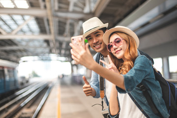 Happy Asian couple traveler holding a mobile phone in station and waiting for train in vacation...
